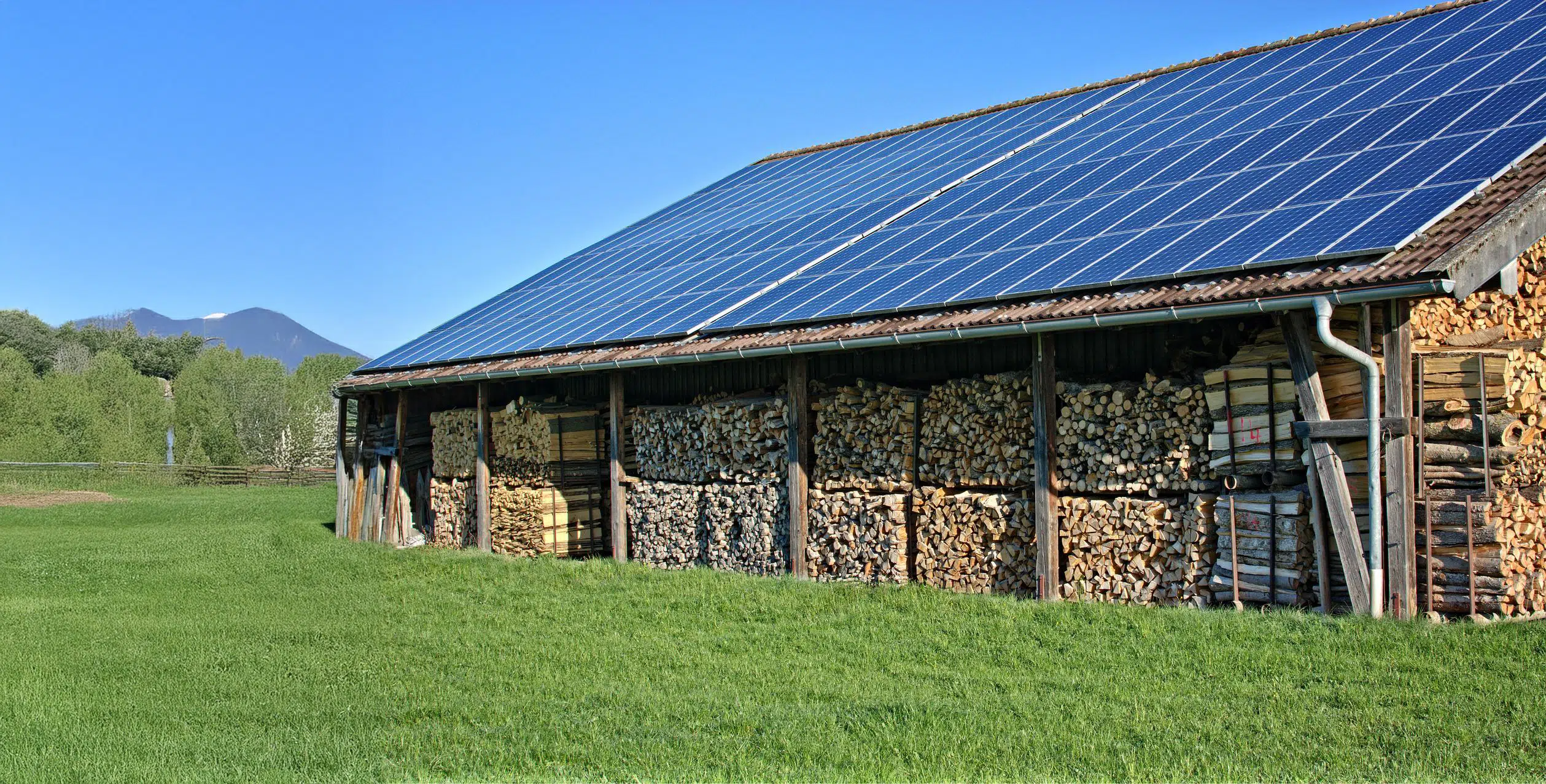 Een houtopslag onder een dak met zonnepanelen, omgeven door groen gras en met bergen op de achtergrond.